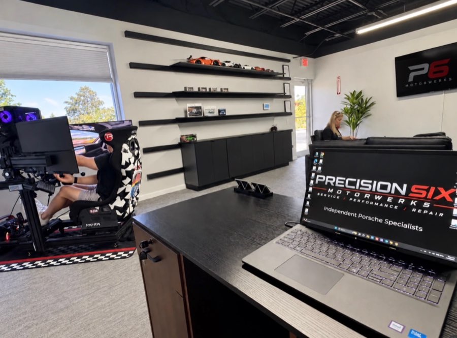 Customer lounge with a racing simulator, model cars on the shelves, and the front desk laptop showing the Precision Six logo