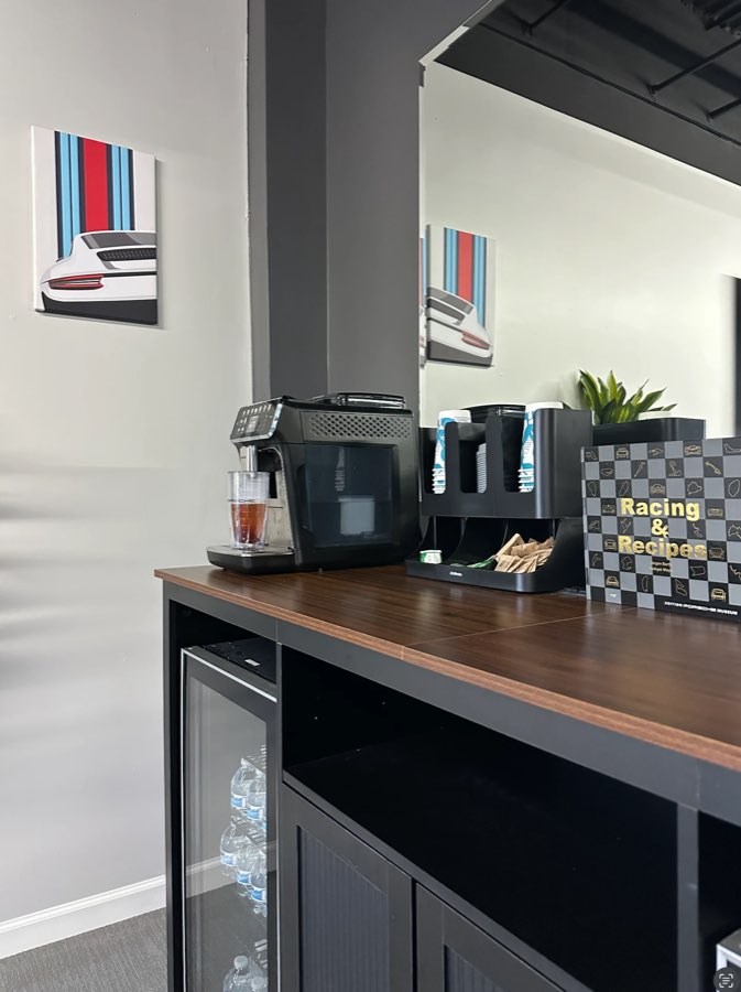 Coffee bar with an espresso machine, a drinks fridge, and a Porsche Museum cookbook, under Martini stripe Porsche art