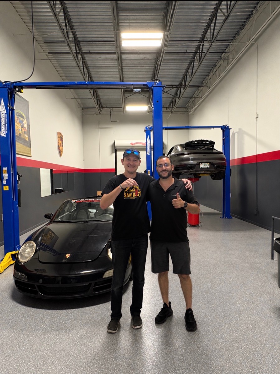 Two members of the P6 team smiling in the shop, one holding up a set of keys and the other giving a thumbs up, in front of a black 911 with a grey 911 on a lift behind them